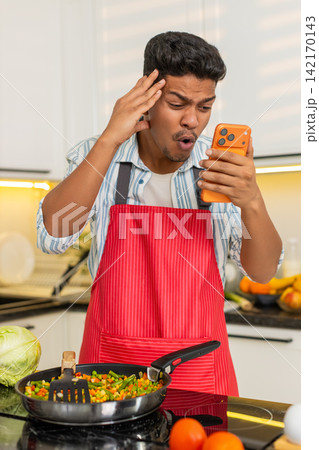 Indian man reading bad smartphone message while frying vegetables in kitchen for healthy meal prep Indian man reading bad smartphone message while frying vegetables in kitchen for healthy meal prep 142170143