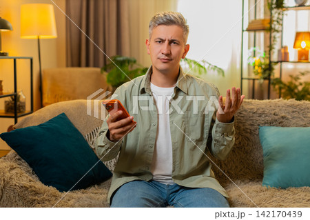 Middle-aged man on home sofa upset after smartphone money charge and online game loss with bad news Middle-aged man on home sofa upset after smartphone money charge and online game loss with bad news 142170439