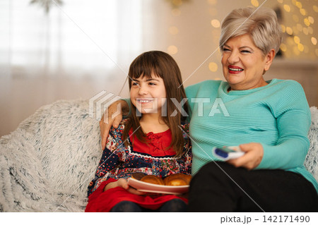 Grandmother granddaughter watching TV together laughing eating snacks 142171490