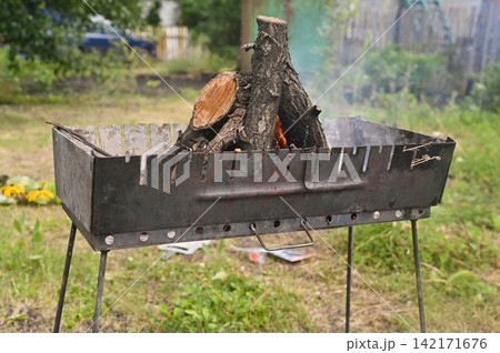 Grill with firewood starting to burn in backyard. Metal grill holding stacked logs with small flame and light smoke, outdoor barbecue equipment on grass, rustic cooking process, summer lifestyle, 142171676
