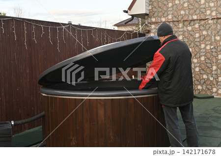 Man opens black cover of wooden outdoor jacuzzi. Adult man prepares round jacuzzi for relaxation in backyard. Wood fired bath stands near fence on autumn day. Garden spa equipment looks ready. Man opens black cover of wooden outdoor jacuzzi. Adult man prepares round jacuzzi for relaxation in backyard. Wood fired bath stands near fence on autumn day. Garden spa equipment looks ready. 142171678