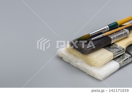 Collection of diverse painting brushes including wide flat brushes and fine detail watercolor tools stacked together on a minimalist gray table background with ample negative space for design copy. 142171839