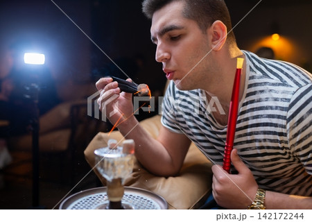 Man blowing on a hot charcoal cube with tongs Man blowing on a hot charcoal cube with tongs 142172244