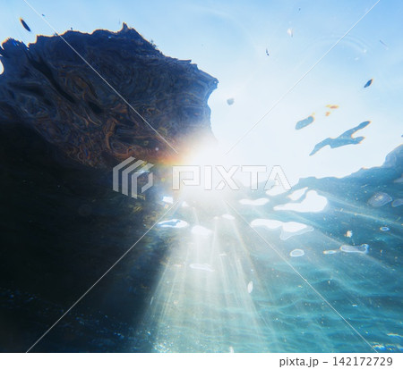 Underwater photography captures bright sun rays piercing through clear ocean water illuminating a dark rocky ledge Underwater photography captures bright sun rays piercing through clear ocean water illuminating a dark rocky ledge 142172729