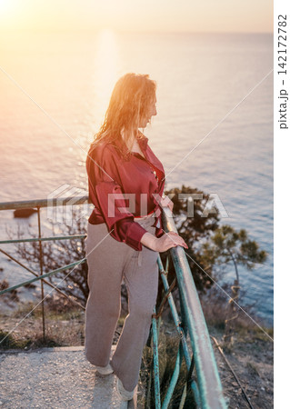 Woman sunset ocean looking at the sea from a balcony at dusk with golden hour light reflecting on the water Woman sunset ocean looking at the sea from a balcony at dusk with golden hour light reflecting on the water 142172782