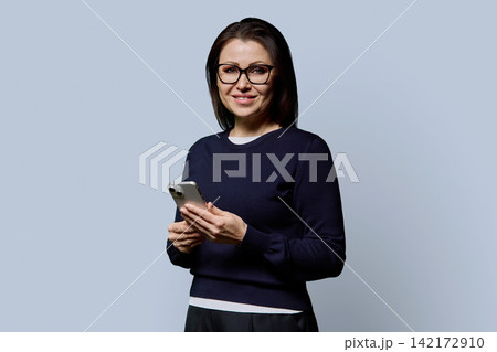 Smiling 40s mature woman in glasses holding smartphone on grey background 142172910