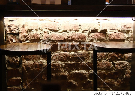 Rustic wooden bar stools resting against a glowing brick wall at night 142173466