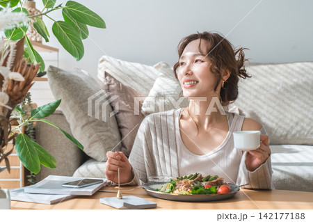 リビングテーブルで食事をするおしゃれな若いアジア人女性 リビングテーブルで食事をするおしゃれな若いアジア人女性 142177188