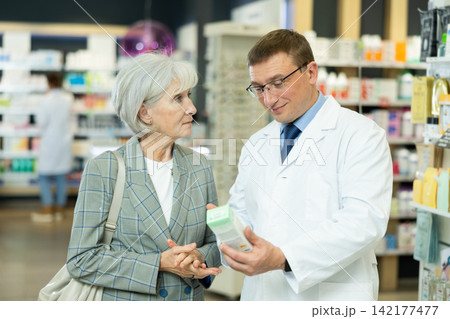 Man apothecary hold package with cream and talks with senior woman client about nuances of care 142177477