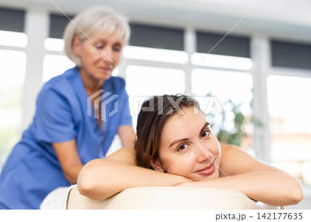 Blurred mature woman actively rubs lower back of young woman during general massage procedure. Blurred mature woman actively rubs lower back of young woman during general massage procedure. 142177635