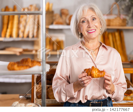 Senior female customer stands with croissant in hands near window of bakery. 142177667