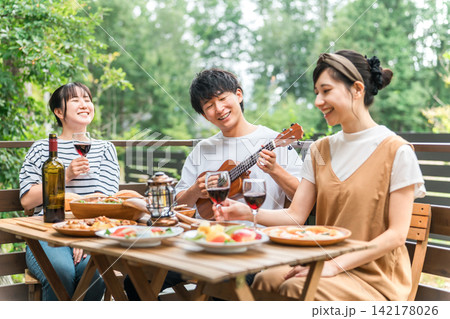 ウッドデッキでウクレレを演奏しながらパーティー料理とお酒を楽しむ男女グループ 142178026