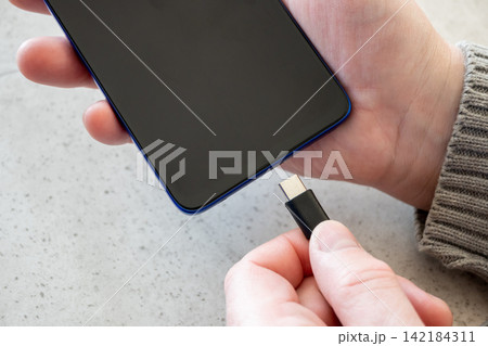 Hand of man girl plugging charger cable into mobile phone on the table. Black cord for smartphone on the stone table. Hand of man girl plugging charger cable into mobile phone on the table. Black cord for smartphone on the stone table. 142184311