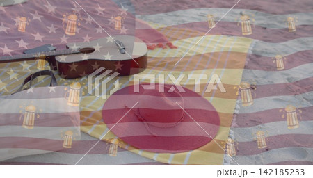 Resting red straw sun hat on yellow-striped towel at sandy beach, with ukulele and flag overlay. Headwear, guitar, picnic, seaside, coastal, outdoors, daylight 142185233