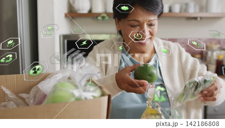 Unpacking senior Indian woman wearing cardigan in kitchen, holding green fruit, box and eco icons Unpacking senior Indian woman wearing cardigan in kitchen, holding green fruit, box and eco icons 142186808