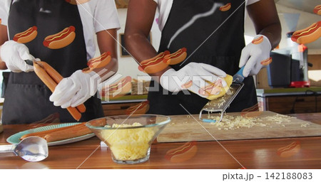 Holding hot dog sausages, staff grating cheese with grater at counter, wearing black aprons, gloves 142188083