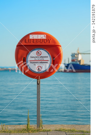 Lifebuoy safety station by sea with cargo ship in background. Red lifebuoy safety station with no swimming and no jumping warning signs on a waterfront by calm blue sea, maritime harbor scene Lifebuoy safety station by sea with cargo ship in background. Red lifebuoy safety station with no swimming and no jumping warning signs on a waterfront by calm blue sea, maritime harbor scene 142191879
