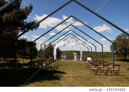 Outdoor wedding ceremony venue with white fabric arch and wooden chairs under metal frame structure in nature. 142193882