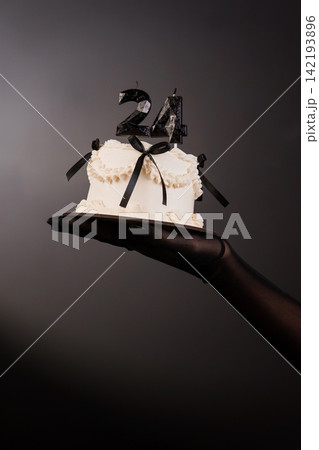 Hand in black glove holding small white cake with black ribbons and number 24 candles on dark grey background. Hand in black glove holding small white cake with black ribbons and number 24 candles on dark grey background. 142193896