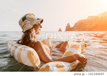 Woman Floating Inflatable Ocean Summer - A woman wearing a hat and sunglasses is floating on an inflatable raft in the ocean, enjoying the summer weather and the view of the cliffs in the distance. 142194097