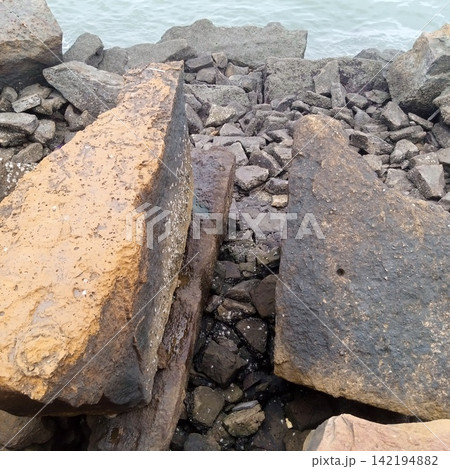 Two Massive Angular Boulders Separated By A Narrow Gap Filled With Crushed Gravel On A Rugged Coastal Shoreline Breakwater Two Massive Angular Boulders Separated By A Narrow Gap Filled With Crushed Gravel On A Rugged Coastal Shoreline Breakwater 142194882