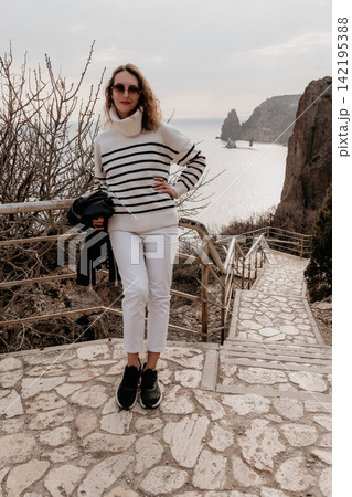 Woman coastline fashion stylish traveler stands on stone steps overlooking scenic ocean cliffs wearing striped sweater and sunglasses 142195388