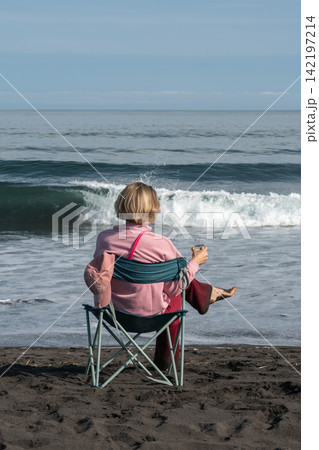 Woman relaxes by Pacific Ocean with glass of champagne while seated on black volcanic sand in Kamchatka. Travel, coastal recreation, peaceful lifestyle, outdoor leisure, solitude, summer vacation. 142197214