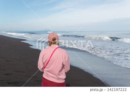 Woman looks toward Pacific Ocean while standing on black volcanic shore at Khalaktyrsky Beach, Kamchatka. Travel, nature tourism, coastal lifestyle, contemplation, solitude, summer vacation. Woman looks toward Pacific Ocean while standing on black volcanic shore at Khalaktyrsky Beach, Kamchatka. Travel, nature tourism, coastal lifestyle, contemplation, solitude, summer vacation. 142197222