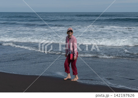 Woman stands barefoot on black volcanic shore with breaking Pacific Ocean waves behind her in Kamchatka. Travel, nature tourism, coastal lifestyle, relaxation, freedom, summer vacation. Woman stands barefoot on black volcanic shore with breaking Pacific Ocean waves behind her in Kamchatka. Travel, nature tourism, coastal lifestyle, relaxation, freedom, summer vacation. 142197626