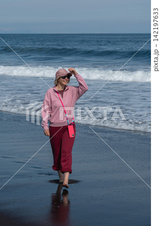 Woman in pink clothes stands barefoot on wet black volcanic sand beside Pacific Ocean waves in Kamchatka. Travel, coastal lifestyle, relaxation, nature tourism, freedom, summer vacation. 142197633
