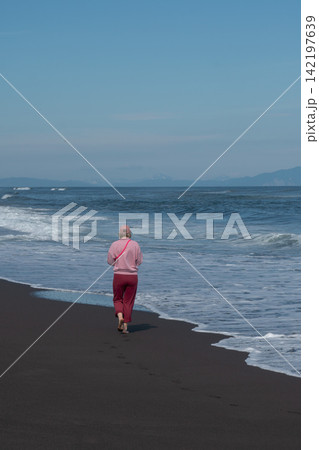 Woman walks along black volcanic shoreline facing Pacific Ocean waves in Kamchatka. Travel, coastal lifestyle, nature tourism, solitude, exploration, outdoor recreation, summer vacation. 142197639