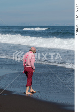 Barefoot woman walks by Pacific waves on black sand at Khalaktyrsky Beach, Kamchatka, Russia. Travel, coastal lifestyle, nature tourism, solitude, summer vacation, outdoor recreation. 142197747