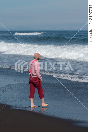 Woman walks barefoot beside Pacific surf on black volcanic beach at Khalaktyrsky Beach, Kamchatka, Russia. Travel, nature tourism, coastal lifestyle, freedom, summer vacation, outdoor recreation. 142197755