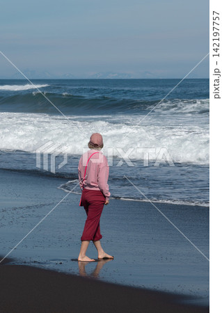 Woman walks barefoot along black volcanic shore beside Pacific Ocean at Khalaktyrsky Beach, Kamchatka, Russia. Travel, nature tourism, coastal lifestyle, solitude, outdoor recreation, summer vacation. 142197757