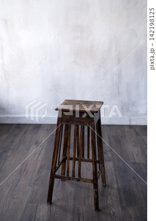 Vintage wooden stool standing on dark hardwood floor against rustic white concrete wall in minimal interior space Vintage wooden stool standing on dark hardwood floor against rustic white concrete wall in minimal interior space 142198125