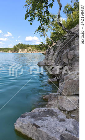 Turquoise quarry lake bordered by rugged rocky cliffs and green forest under a bright summer sky, with clear rippling water creating a scenic natural landscape for travel and outdoor concepts. Turquoise quarry lake bordered by rugged rocky cliffs and green forest under a bright summer sky, with clear rippling water creating a scenic natural landscape for travel and outdoor concepts. 142199487