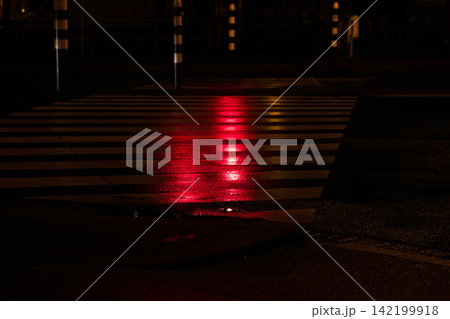 Night city street with a zebra crossing illuminated by reflections from red stop signals on wet asphalt. Pedestrian crosswalk with traffic lights. Road safety in the darkness, nightlife concept Night city street with a zebra crossing illuminated by reflections from red stop signals on wet asphalt. Pedestrian crosswalk with traffic lights. Road safety in the darkness, nightlife concept 142199918