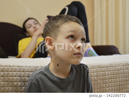 A young boy in a gray T-shirt is sitting on the edge of a bed or mattress. A young boy in a gray T-shirt is sitting on the edge of a bed or mattress. 142200772