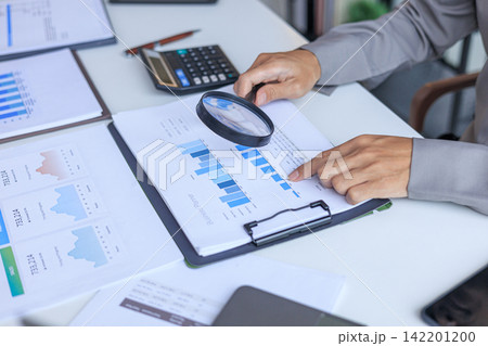 Person using a magnifying glass to review financial and business growth bar charts on a white desk. 142201200