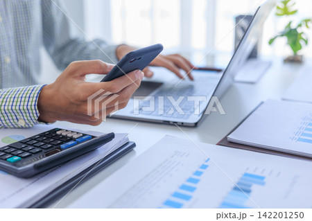 Businessman working in office using smartphone and typing on laptop with graph data and calculator at desk. Businessman working in office using smartphone and typing on laptop with graph data and calculator at desk. 142201250