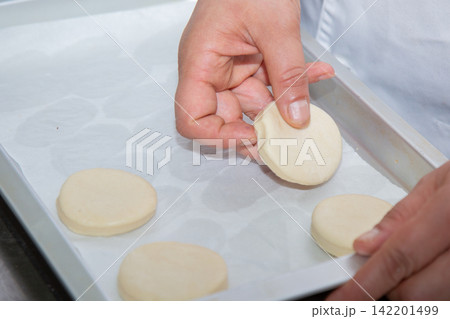Hands placing round dough pieces on parchment lined baking tray in close up kitchen preparation scene with soft natural light, careful shaping, baking process, homemade pastry making 142201499