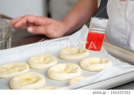 Cook brushing oil onto shaped dough rounds on baking tray before baking in bright kitchen, close view of pastry preparation with metal tray, parchment paper, hand, brush, and soft natural light 142201501