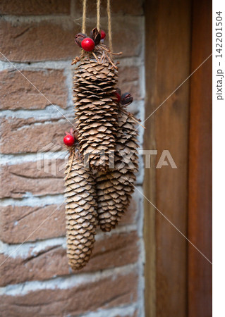Hanging pinecone decoration with red berries displayed against rustic brick wall, showcasing natural texture, warm brown tones, and handcrafted seasonal ornament detail in soft indoor light Hanging pinecone decoration with red berries displayed against rustic brick wall, showcasing natural texture, warm brown tones, and handcrafted seasonal ornament detail in soft indoor light 142201504