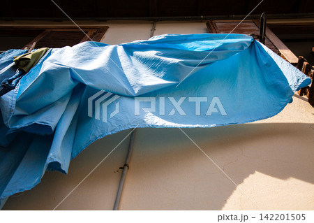 Blue laundry hanging from balcony clothesline beside sunlit wall, viewed from low angle with wooden shutters, soft fabric folds, warm daylight and urban domestic atmosphere 142201505