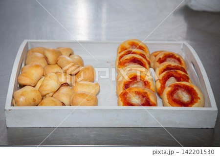 Tray of freshly baked bread rolls and small pizza rounds arranged on stainless steel counter with warm golden crusts, simple bakery food preparation scene in bright kitchen 142201530