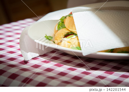 Fresh bread rolls on white plate beneath napkin, warm sunlight over red and white checkered tablecloth creating rustic breakfast dining mood in cozy indoor setting Fresh bread rolls on white plate beneath napkin, warm sunlight over red and white checkered tablecloth creating rustic breakfast dining mood in cozy indoor setting 142201544