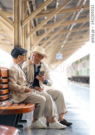 Two senior LGBTQ men in cream suits share laughter on train station bench, one holding phone, one with camera 142205032