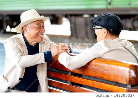 Two senior LGBTQ men share warm handshake on wooden bench at train station, one in straw hat, one in cap Two senior LGBTQ men share warm handshake on wooden bench at train station, one in straw hat, one in cap 142205037