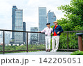 Two senior LGBTQ men stand on modern balcony, one holds camera as they gaze at city skyline through lush greenery 142205051