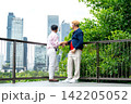 Two senior LGBTQ men converse on modern balcony railing, city skyline and lush greenery behind 142205052